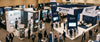 An elevated view of a professional trade show floor featuring two different-sized exhibit booths side by side, surrounded by softly blurred attendees under warm venue lighting.