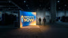 A glowing backlit trade show display stands out brilliantly against the dim, shadowy interior of a large convention hall, attracting attendees from across the floor.