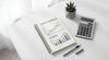 Overhead flat-lay of a notebook with charts, a calculator, and a pen on a clean white desk, evoking strategic business planning and ROI analysis.