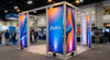 A modern trade show booth with a sturdy aluminum frame fitted with fresh, vibrant fabric graphic panels glowing under warm exhibition hall lighting.