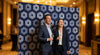 Two professionals posing in front of a professional step-and-repeat banner at a branded corporate event.