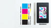 Flat-lay overhead view of large-format print materials, color swatches, a ruler, and a design tablet arranged on a clean white surface.
