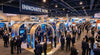 A busy trade show exhibition hall with a prominently designed booth in the foreground attracting a crowd of professional attendees amid rows of competing exhibits.